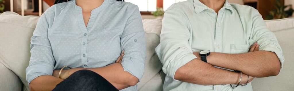 couple sits on a couch each with their arms crossed. They are experiencing mismatched libido or desire discrepancy. They work with a sex therapist in Cincinnati, OH at Emma Schmidt and Associates